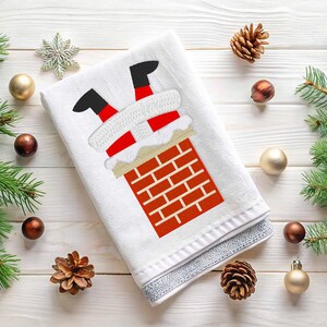 May include: White kitchen towel with an embroidered design of Santa Claus getting stuck in a chimney. The design features Santa's legs and feet sticking out of the chimney, with a red and white Santa suit and a white beard. The chimney is made of red bricks.