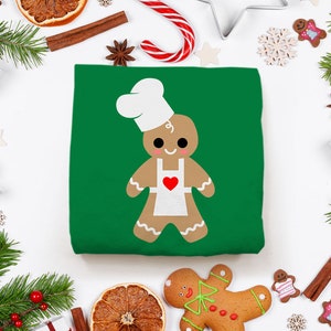 May include: A green t-shirt with a gingerbread man wearing a chef's hat and apron. The gingerbread man is smiling and has a red heart on his apron. The t-shirt is surrounded by Christmas decorations, including pine branches, ornaments, and candy canes.