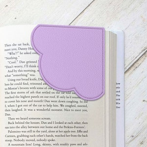 Può includere: Un segnalibro viola con una forma unica, con un bordo cucito e una superficie testurizzata. Il segnalibro è posizionato su un libro aperto.