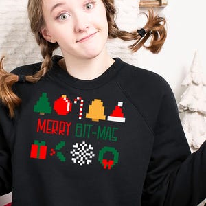 Puede incluir: Una sudadera negra con un diseño navideño pixelado que dice "Merry Bit-Mas". El diseño incluye un árbol de Navidad, adornos, bastones de caramelo, campanas y un gorro de Papá Noel.
