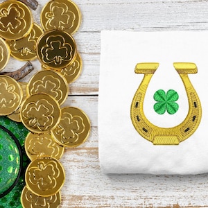 Puede incluir: Una tela blanca con una herradura dorada y un trébol de cuatro hojas verde bordado en ella. Hay monedas de oro con tréboles verdes en el fondo.