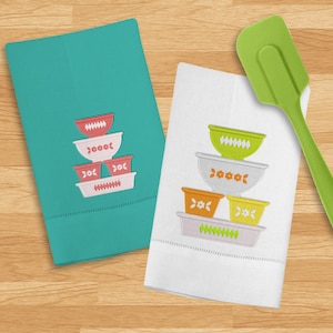 May include: Two white kitchen towels with embroidered images of stacked bowls. The bowls are in various colors, including red, orange, yellow, and green. A green spatula is on the right side of the image.