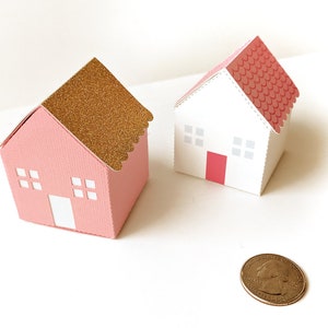May include: Two small paper houses, one pink with a gold glitter roof and the other white with a pink roof. Both houses have white windows and a red door.