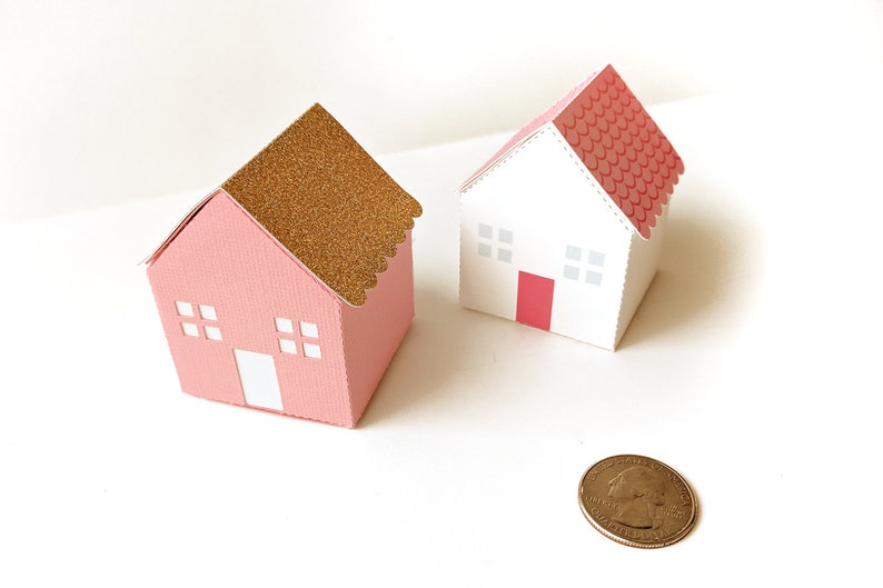 May include: Two small paper houses, one pink with a gold glitter roof and the other white with a pink roof. Both houses have white windows and a red door.