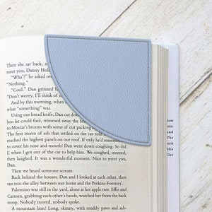 May include: A light blue leather bookmark with a rounded corner. The bookmark is placed on a book with white pages and black text.