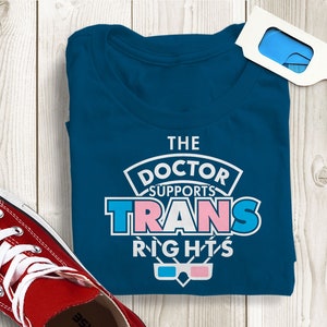 Pode incluir: Camiseta azul-petróleo com o texto "THE DOCTOR SUPPORTS TRANS RIGHTS" em branco, rosa e azul. Um par de óculos 3D e um tênis vermelho também são visíveis.