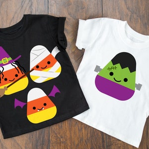 May include: Two black and white t-shirts with candy corn designs. One shirt features a candy corn witch with a broom and a candy corn mummy. The other shirt features a candy corn Frankenstein.