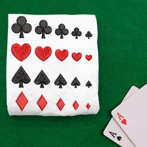 May include: A white fabric with embroidered playing card suits: clubs, hearts, spades, and diamonds. Two playing cards with the Ace of Hearts and Ace of Spades are on a green surface.