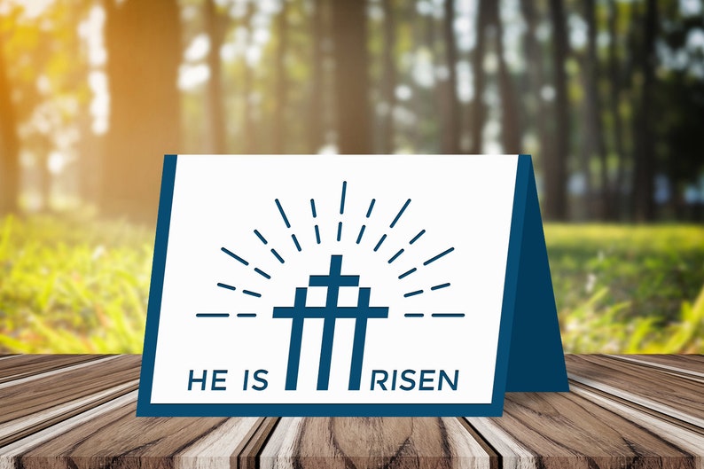 May include: A white card with a blue graphic of three crosses with a sunburst behind them. The text "HE IS RISEN" is printed below the graphic. The card is standing on a wooden table.