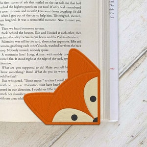 May include: Orange fox-shaped bookmark made of fabric, placed on the corner of a book. The bookmark features a fox face with black eyes and a cream-colored muzzle. The book's pages are visible, with text printed on them.