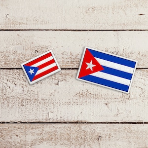 May include: Two small flags on a white wooden background. The flag on the left is the flag of Puerto Rico, and the flag on the right is the flag of Cuba.