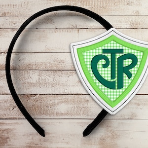 May include: A black headband with a shield-shaped emblem. The emblem features a green and white gingham pattern, a green border, and the letters "CTR" in a stylized font. The design is set against a light wood background.