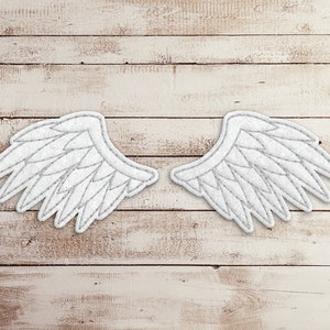 May include: A pair of white felt angel wings with a gray outline, against a light brown wood background.
