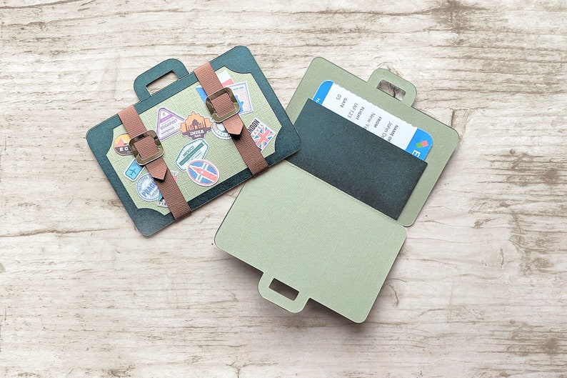 May include: A green and brown paper craft suitcase with a ticket pocket and a handle. The suitcase is decorated with travel stickers and has a brown leather strap.