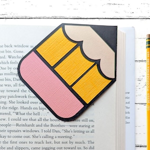 Könnte beinhalten: Ein rosa und gelbes Lesezeichen in Form eines Bleistifts mit schwarzem Umriss. Das Lesezeichen ist aus Papier und hat eine strukturierte Oberfläche.