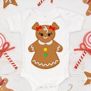 Puede incluir: Un body blanco para bebé con un diseño de hombre de jengibre. El hombre de jengibre es marrón con detalles de glaseado rojo y blanco. El body está rodeado de bastones de caramelo y galletas de jengibre.