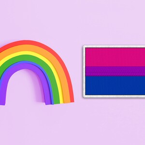 Pode incluir: Um arco-íris e uma bandeira do orgulho bissexual com listras rosa, roxas e azuis sobre um fundo roxo claro.