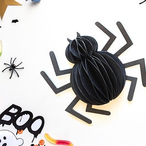 May include: Black paper spider with a 3D layered body and black paper legs. The spider is on a white background with other Halloween decorations, including a pumpkin, a ghost, and candy.