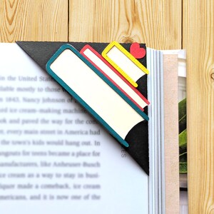 May include: A black, teal, red and yellow layered book-shaped bookmark with a red heart on the top layer. The bookmark is placed in a book.