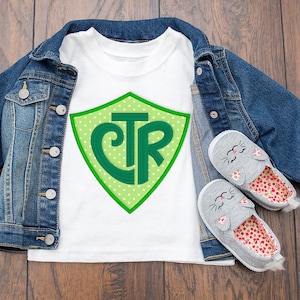 May include: White t-shirt with a green shield design featuring the letters "CTR" on a white background with green polka dots. The shirt is laid out on a wooden surface with a denim jacket and a pair of gray cat-themed shoes.