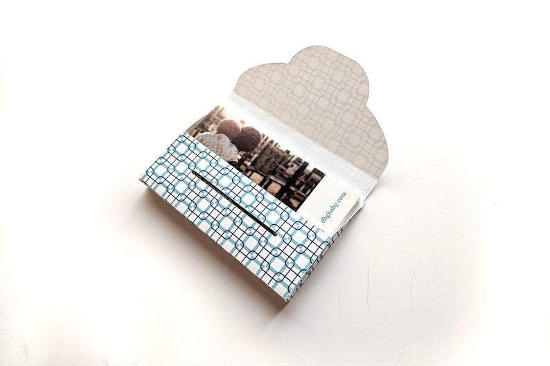 May include: A white and blue patterned box with a photo of two people inside. The box has a flap that opens to reveal the photo. The text "digibaby.com" is printed on the box.