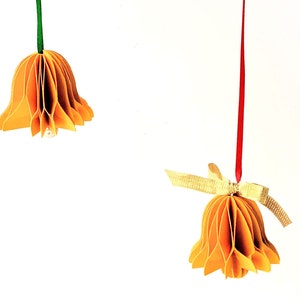 May include: Two orange paper bells hanging from strings. The bell on the left has a green string and the bell on the right has a red string with a gold ribbon.