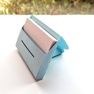 May include: A light blue cardboard business card holder with a slot for cards. The holder is open and has a stack of white business cards inside.