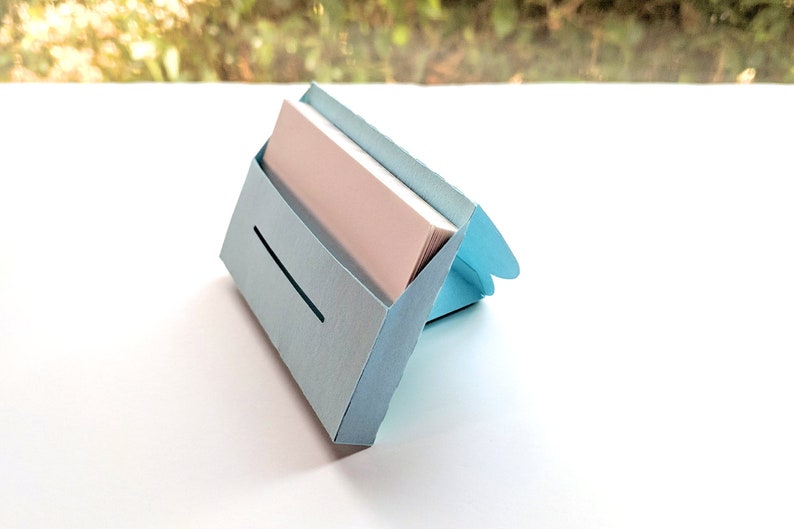 May include: A light blue cardboard business card holder with a slot for cards. The holder is open and has a stack of white business cards inside.
