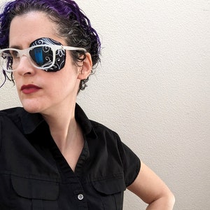 May include: An eye patch with a black and white pattern is attached to clear-framed glasses. The eye patch covers the left eye. The person is wearing a black button-down shirt.