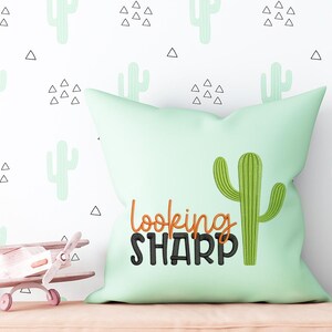 Puede incluir: Una almohada verde con un cactus y el texto "looking sharp" bordado en ella. La almohada está sobre un fondo blanco con un patrón repetitivo de pequeños cactus verdes y triángulos negros.