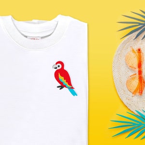 Puede incluir: Camiseta blanca con un loro rojo, amarillo y azul bordado en la parte delantera. El loro está mirando hacia la izquierda y tiene un pico azul. La camiseta está sobre un fondo amarillo.