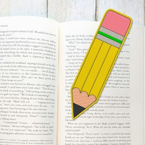 Peut inclure: Un marque-page en forme de crayon jaune avec une gomme rose et une bande verte. Le marque-page est en cuir et a une pointe en forme de cœur. Le marque-page est placé sur un livre ouvert.