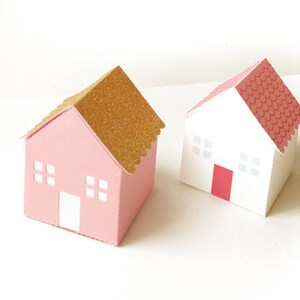 May include: Two small paper houses, one pink with a gold glitter roof and one white with a red roof. Both houses have white windows and a red door.