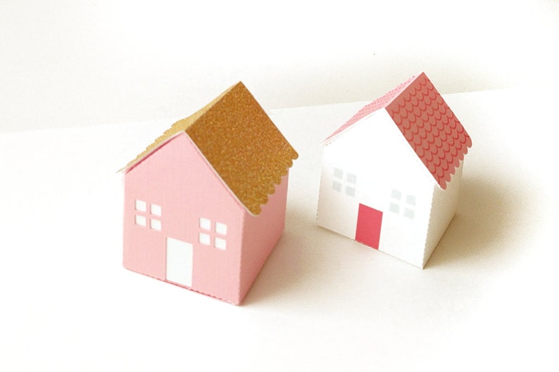 May include: Two small paper houses, one pink with a gold glitter roof and one white with a red roof. Both houses have white windows and a red door.