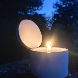 Puede incluir: Una vela blanca encendida en un soporte redondo con tapa, sobre un cielo azul oscuro y árboles. La llama de la vela brilla intensamente, iluminando el interior del soporte. La escena evoca una sensación de tranquilidad.