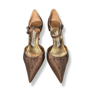 May include: A pair of brown patterned high heels with pointed toes. The shoes feature ankle straps and a gold-colored interior. The brand name "MANOLO BLAHNIK" is visible on the inner sole.