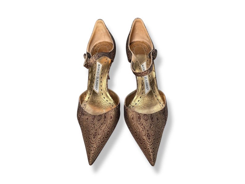 May include: A pair of brown patterned high heels with pointed toes. The shoes feature ankle straps and a gold-colored interior. The brand name "MANOLO BLAHNIK" is visible on the inner sole.