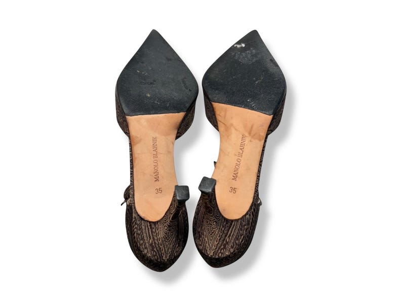 May include: A pair of Manolo Blahnik size 35 heels. The shoes have a pointed toe and a patterned fabric. The soles are black, and the heels have a small, square detail. The shoes are brown and black.