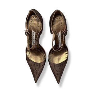 May include: A pair of brown high-heeled shoes with a pointed toe and a strap. The shoes have a patterned design and a gold-coloured interior. The brand name "MANOLO BLAHNIK" is visible on the insole.