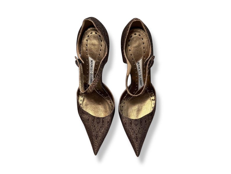 May include: A pair of brown high-heeled shoes with a pointed toe and a strap. The shoes have a patterned design and a gold-coloured interior. The brand name "MANOLO BLAHNIK" is visible on the insole.