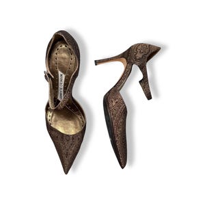 May include: A pair of brown high-heeled shoes with a pointed toe and ankle strap. The shoes feature a paisley pattern and a stiletto heel. The interior of the shoe is gold-toned, and the brand name "MANOLO BLAHNIK" is visible on the insole.