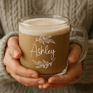 May include: A clear glass mug filled with a creamy drink, with the name "Ashley" in white script and a floral wreath. The mug has a handle and is held by someone wearing a grey jumper.