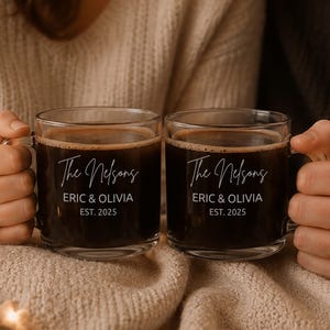 May include: Two clear glass mugs filled with dark coffee, each featuring white printed text that reads The Nelsons, ERIC & OLIVIA, and EST. 2025. The mugs have a simple handle design and are held by two individuals wearing cozy knit sweaters.