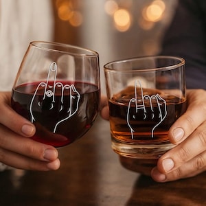 May include: Two clear glasses, one a stemless wine glass and the other a rocks glass, each with a white graphic of a hand giving the middle finger. The wine glass contains red liquid, and the rocks glass contains a brown liquid.