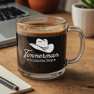 Peut inclure: Une tasse à café en verre transparent remplie de café noir. La tasse présente un dessin blanc d'un chapeau de cowboy et le texte "Timmerman Accessories Shop". La tasse est posée sur une surface en bois à côté d'un ordinateur portable et d'un bloc-notes.