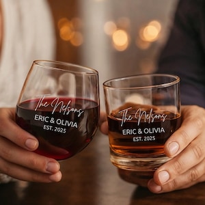 May include: Two clear glass vessels, one stemless wine glass filled with red liquid and one rocks glass filled with amber liquid, both featuring white printed text that reads The Nelsons, ERIC & OLIVIA, and EST. 2025 in a mix of script and sans-serif fonts.