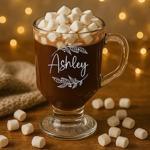 May include: A clear glass mug filled with hot chocolate and topped with marshmallows. The mug has a handle and the name "Ashley" is written on the front in a decorative font. The mug sits on a wooden surface.