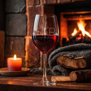 May include: A clear wine glass filled with red wine, featuring the initials "S & R" and a floral design. The name "Sarah Rogers" is printed below the initials. A lit candle and a fireplace with burning logs are in the background.
