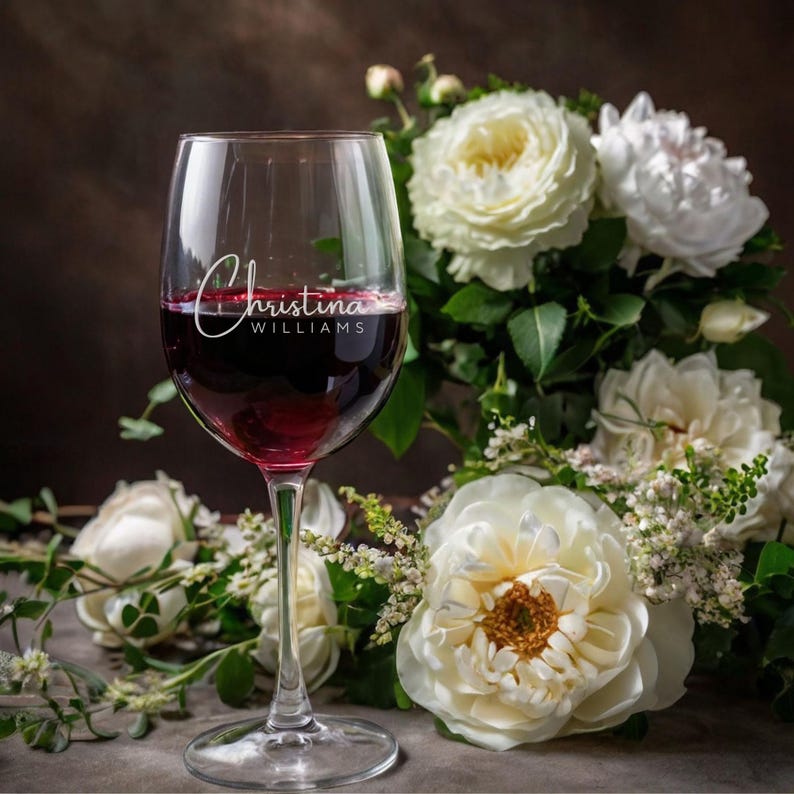 May include: A wine glass filled with red wine, featuring the name "Christina Williams" in elegant script. The glass is set against a backdrop of white roses and greenery, creating a romantic and elegant scene.