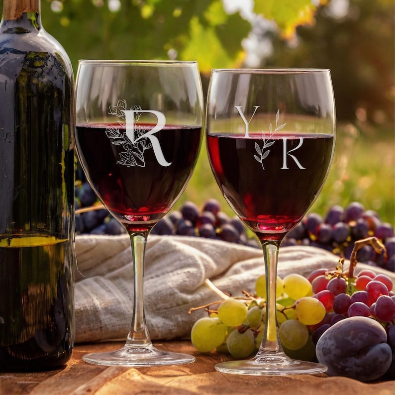 May include: Two wine glasses filled with red wine, each etched with a letter and floral design. One glass has the letter "R", the other has "Y" and "R". A wine bottle is on the left, and grapes are in the background.